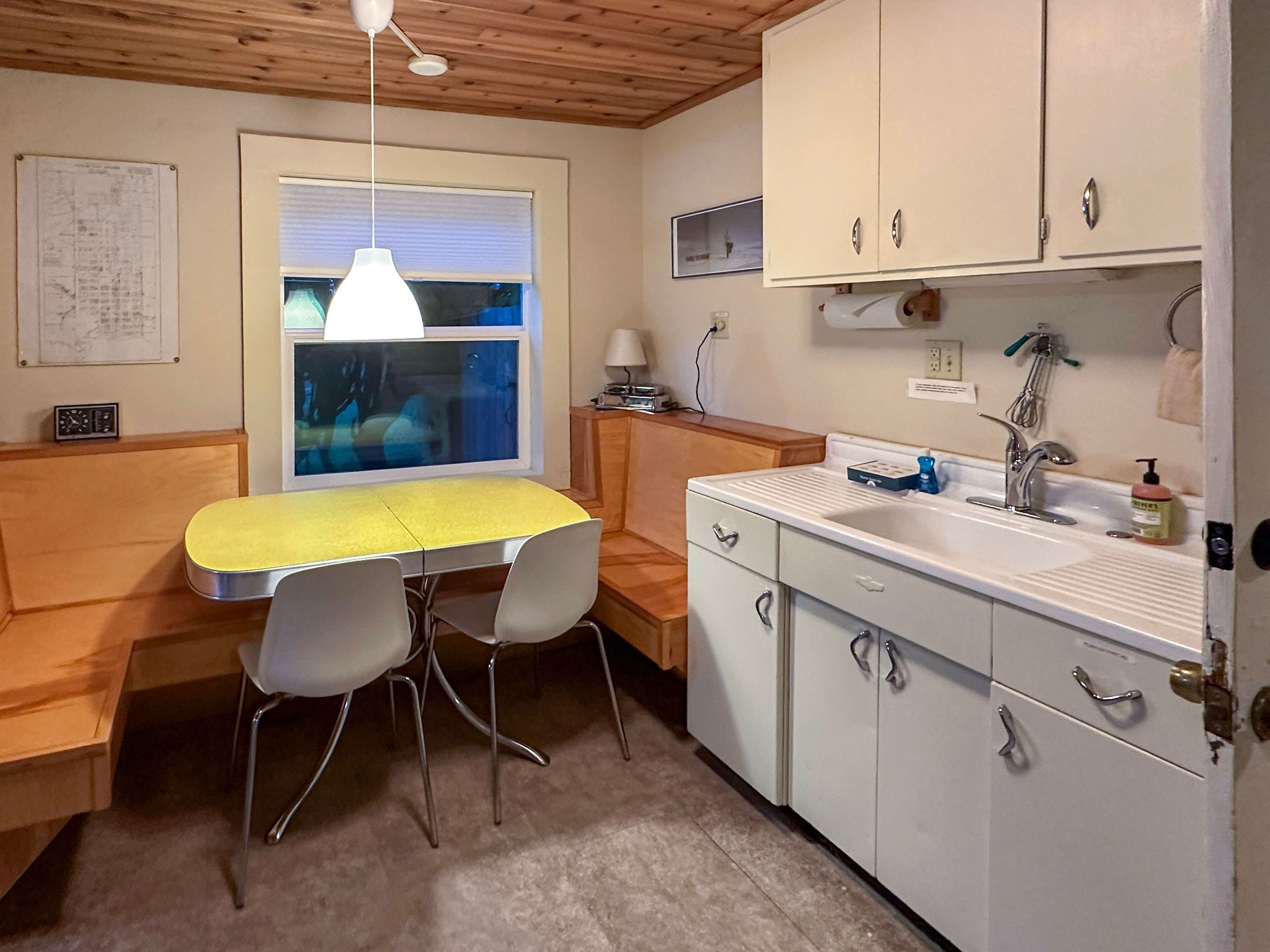 A compact kitchen with a yellow table, two chairs, a window with blinds, a sink, and cabinets.