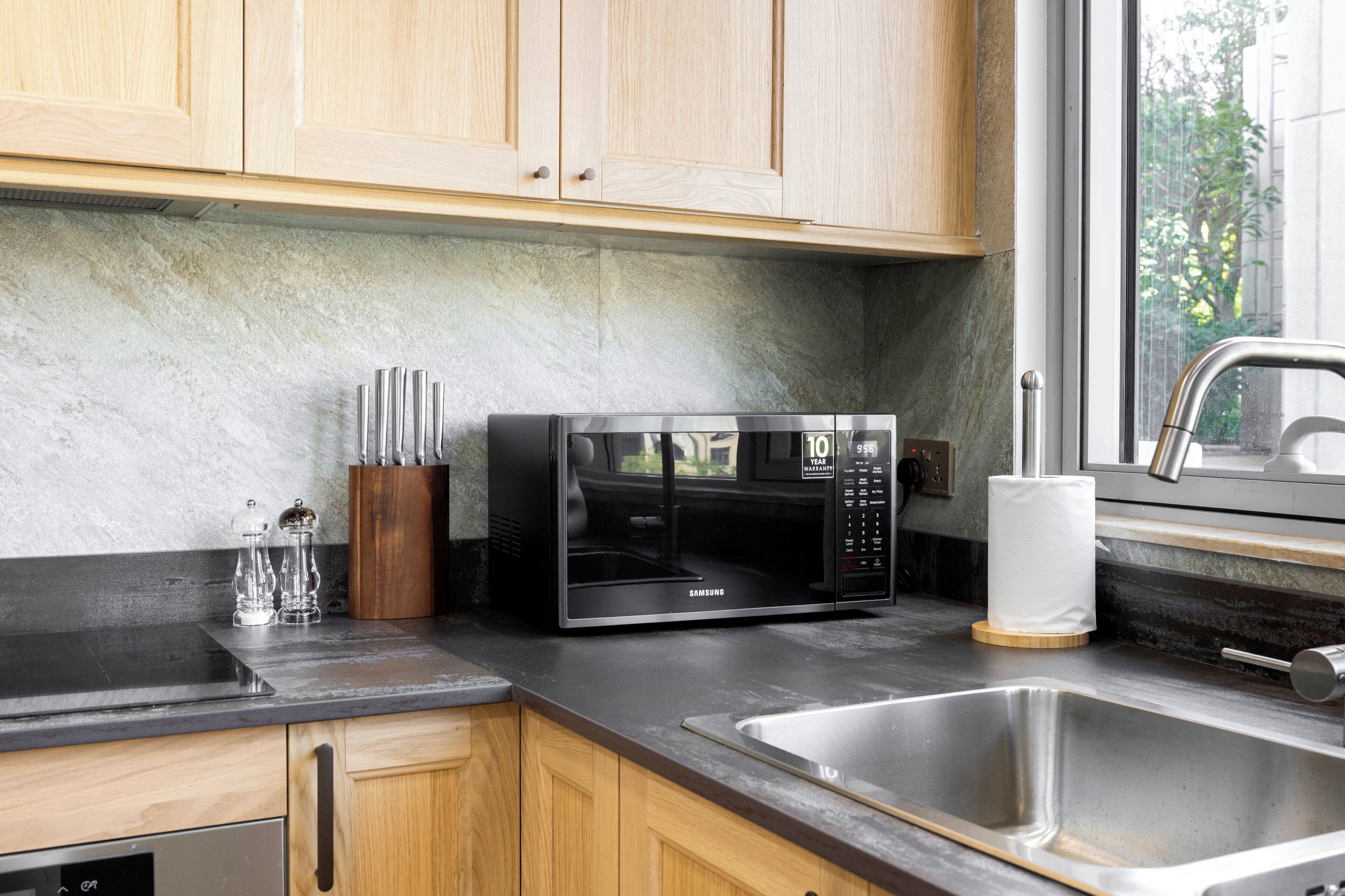 Een moderne keuken met houten kastjes, een zwarte magnetron, een roestvrijstalen spoelbak en een raam met uitzicht op groen.