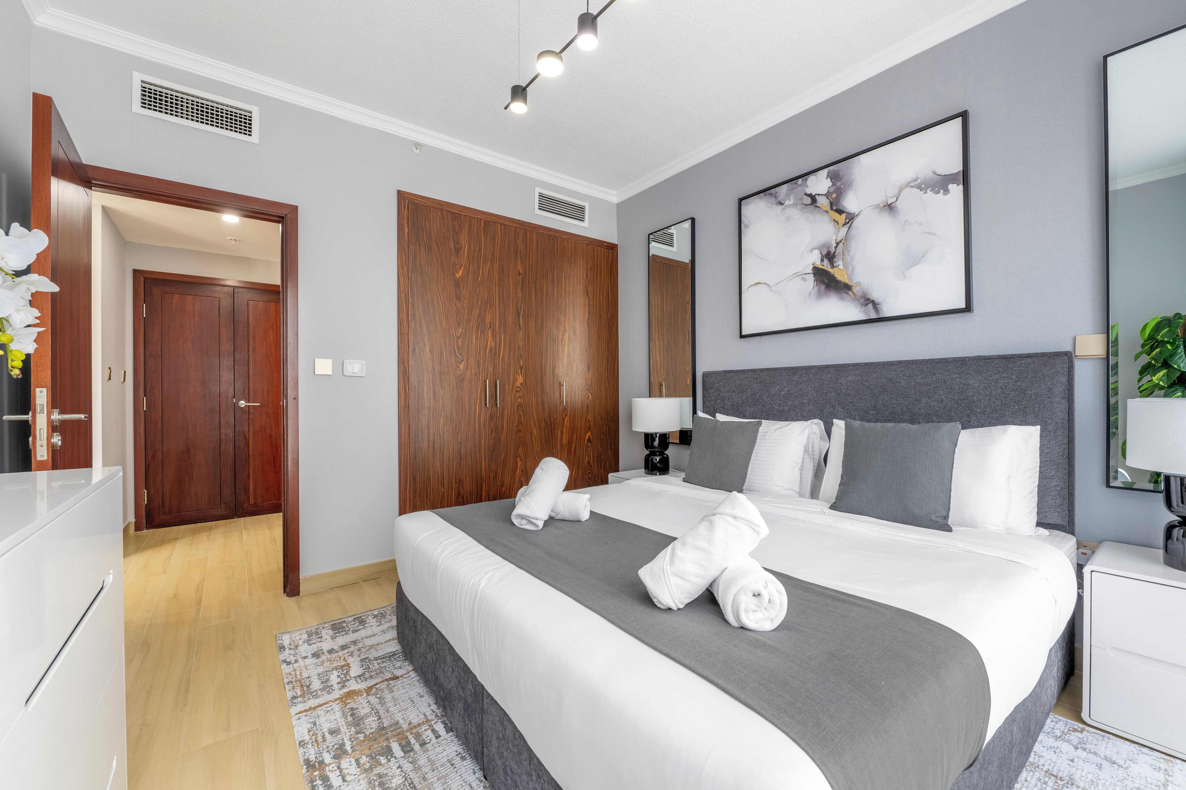 A modern hotel room with a large bed, wooden wardrobe, and a grey headboard.