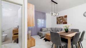 Uma sala de estar moderna com um sofá cinzento, mesa de centro de madeira e cadeiras cinzentas. Uma mesa de jantar com um jogo americano trançado e flores brancas está no centro. Um armário de madeira e uma pia branca são visíveis ao fundo.