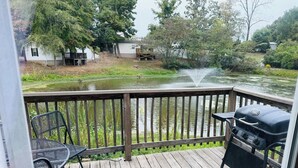 A wooden deck with a grill and chairs, overlooking a pond and houses in the background.