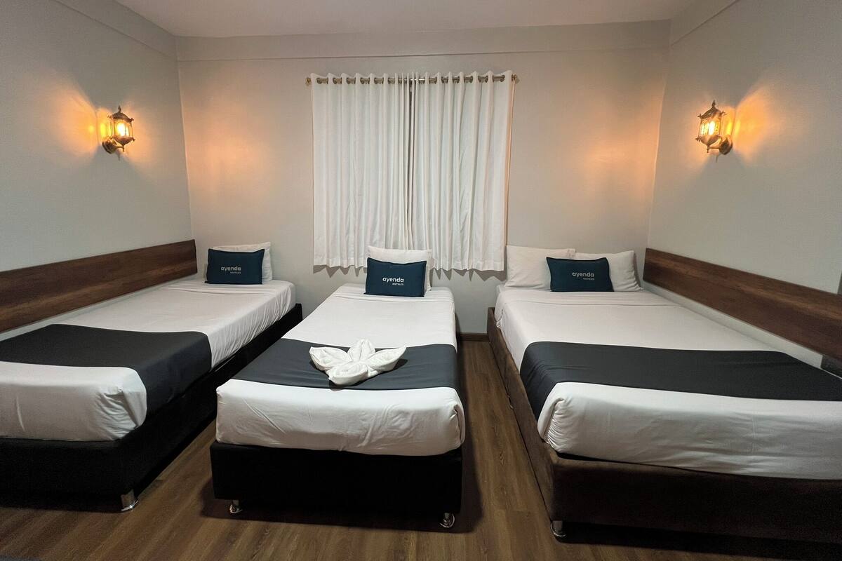 Ein Hotelzimmer mit drei Einzelbetten, jedes mit Kissen und Decke. An beiden Seiten des Fensters befinden sich je zwei Wandleuchten.
