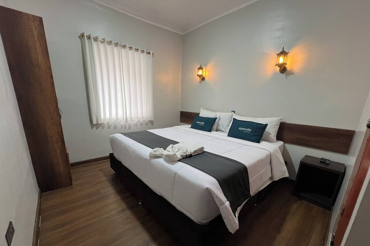 Ein Hotelzimmer mit einem großen Bett, zwei Kissen mit dem Markennamen, einem Nachttisch, einer Fenster mit Vorhängen und Wandleuchten.