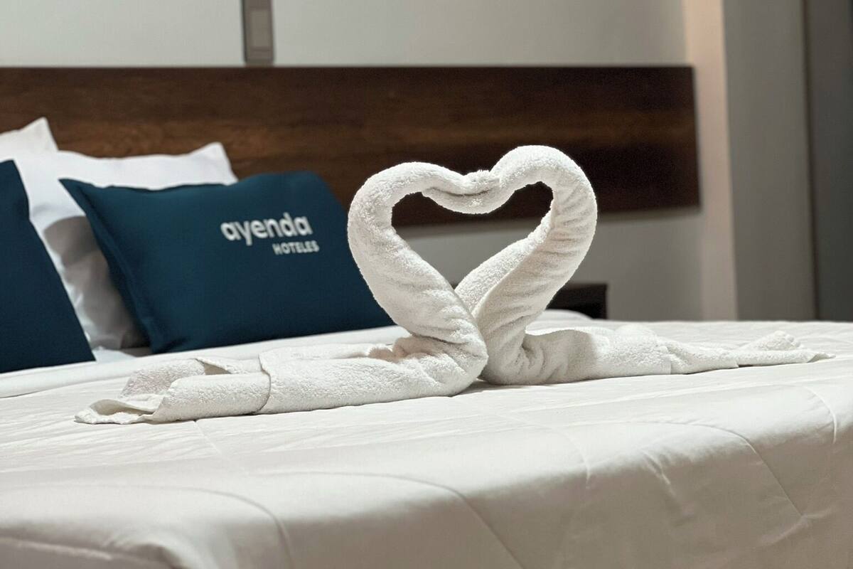 Ein ordentlich bezogenes Bett mit einer herzförmigen Handtuchgestaltung, ein blaues Kissen mit dem Aufdruck "ayenda hotel" und ein türkisblaues Kopfteil.