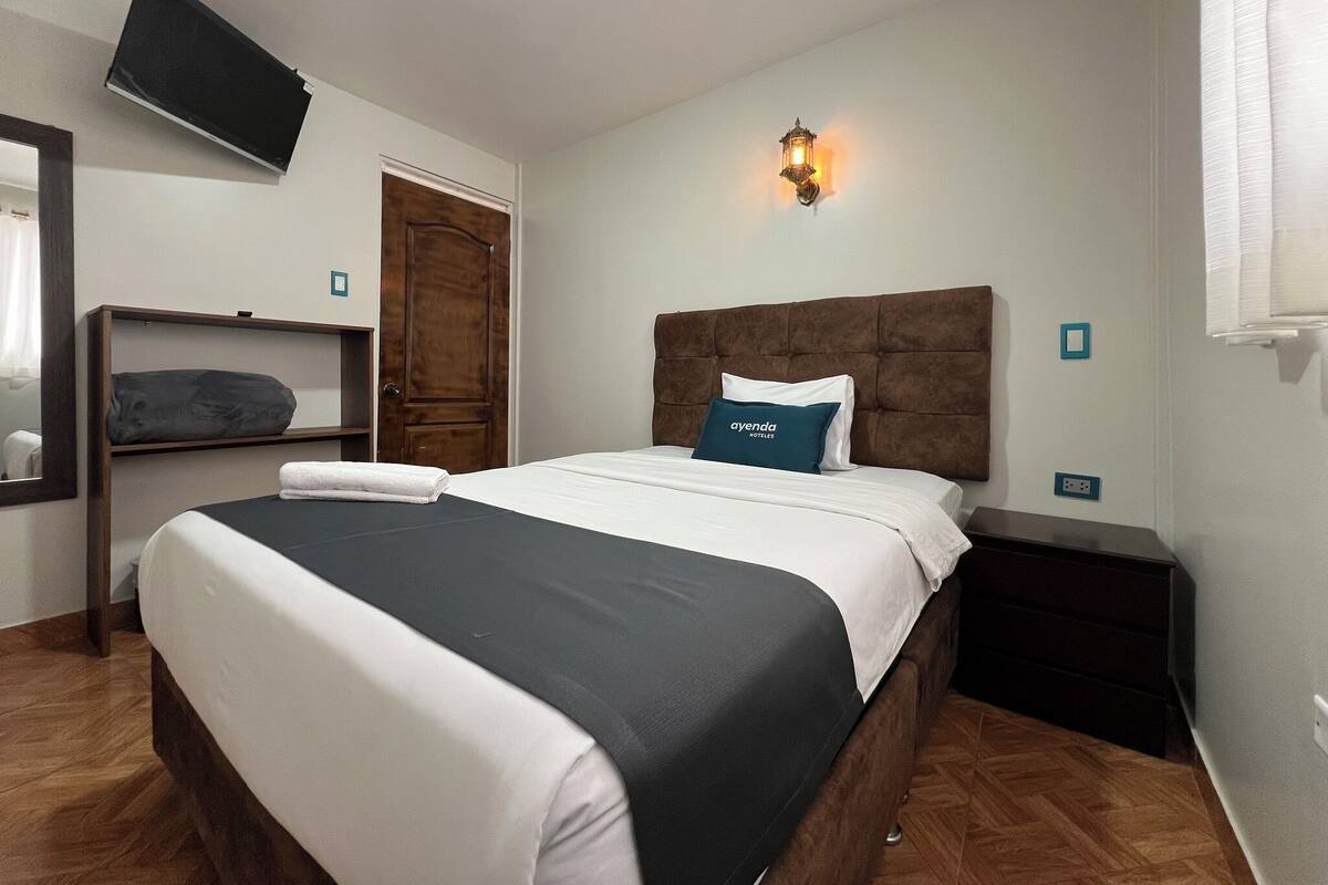Ein Hotelzimmer mit einem großen Bett, einem an der Wand befestigten Fernseher, einer Holztür und einem Nachttisch mit Lampe.