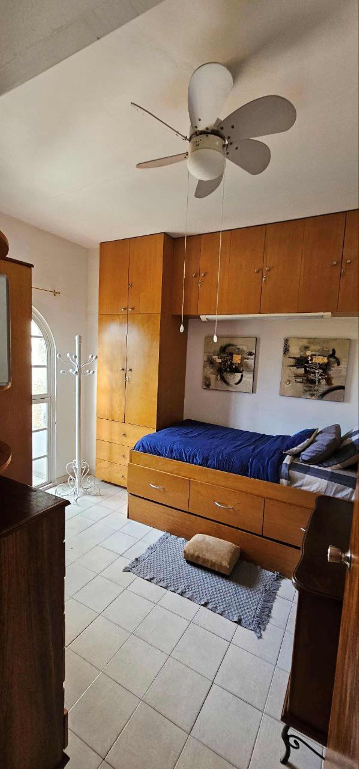 Een slaapkamer met een bed, een houten kledingkast, een plafondventilator en kunst aan de muur.