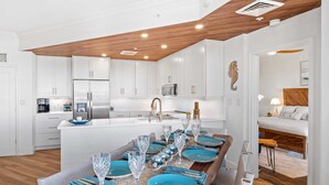 A modern kitchen with white cabinetry, a wooden ceiling, and a dining area set with blue plates and wine glasses. - SeaCrest 2512 Stunning View, Walk to Coligny Shops & Restaurants (Hilton Head Island)