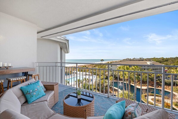 A balcony with a view of the sea, a pool, and a dining area with a table and chairs. - SeaCrest 2512 Stunning View, Walk to Coligny Shops & Restaurants (Hilton Head Island)