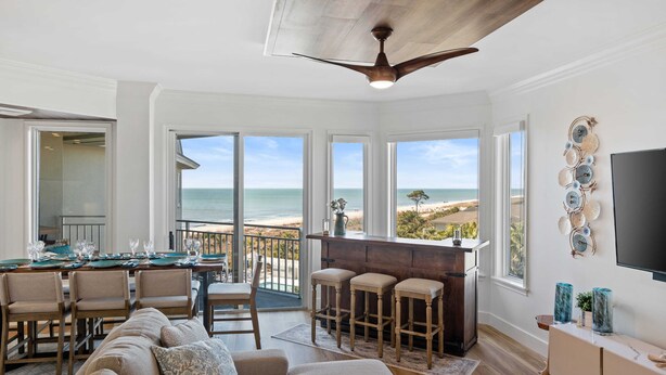 A spacious living room with a large window overlooking the beach, a ceiling fan, and a bar area. - SeaCrest 2512 Stunning View, Walk to Coligny Shops & Restaurants (Hilton Head Island)