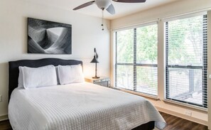 Un dormitorio con una cama, ventilador de techo, mesita de noche, lámpara y ventanas grandes con persianas.