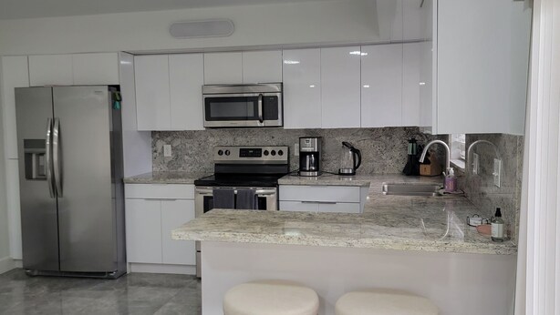 Una cocina moderna con armarios blancos, un refrigerador de acero inoxidable, un microondas, una cafetera y dos taburetes de madera.
