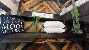 A cozy room with a herringbone-patterned wooden floor, a bed with white pillows, a sign reading "COME IN and stay awhile", and a wall-mounted shelf with a sign and hanging plants.