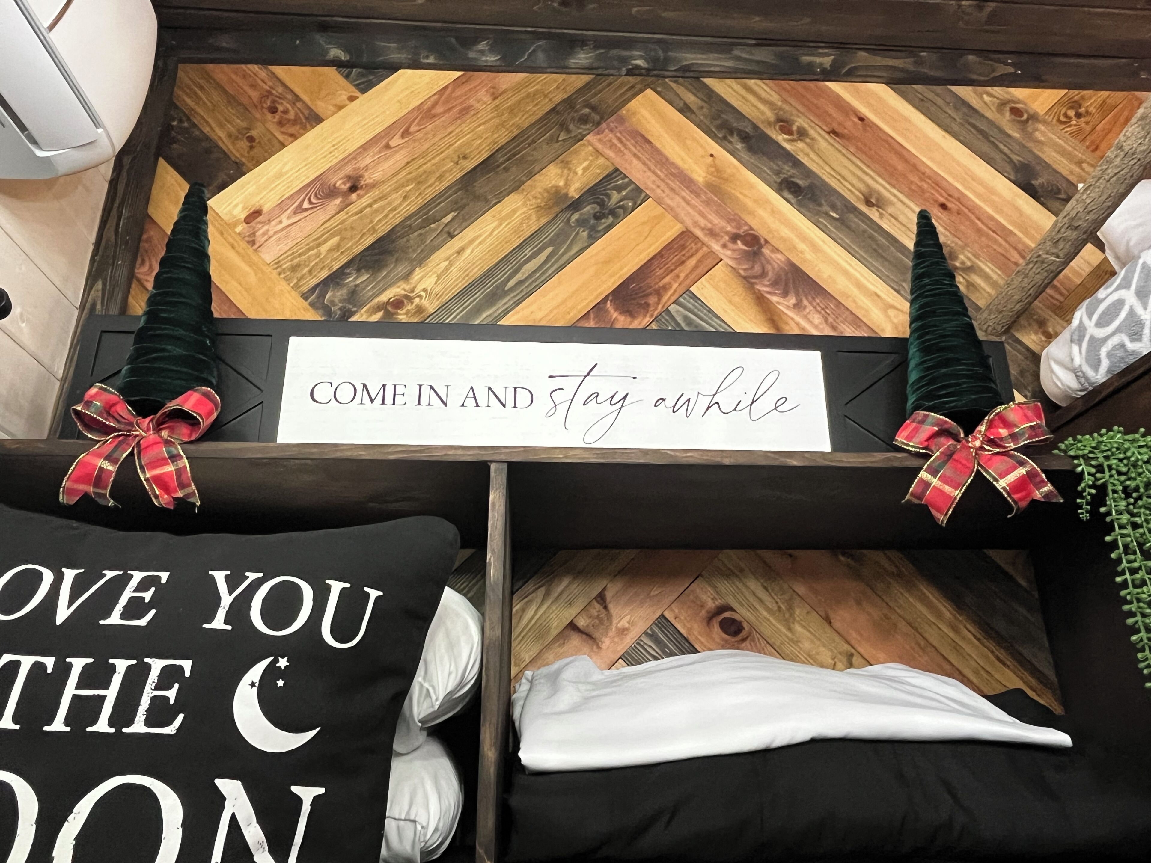 A cozy room with a wooden herringbone floor, a sign that reads "COME IN AND Stay awhile", and two pillows with bows.