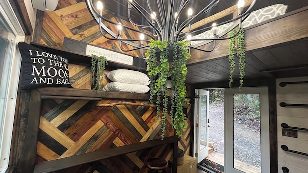 A cozy room with a wooden herringbone floor, a bed with a sign saying "I love you to the moon and back", a hanging plant, and a view of the outdoors through a glass door.