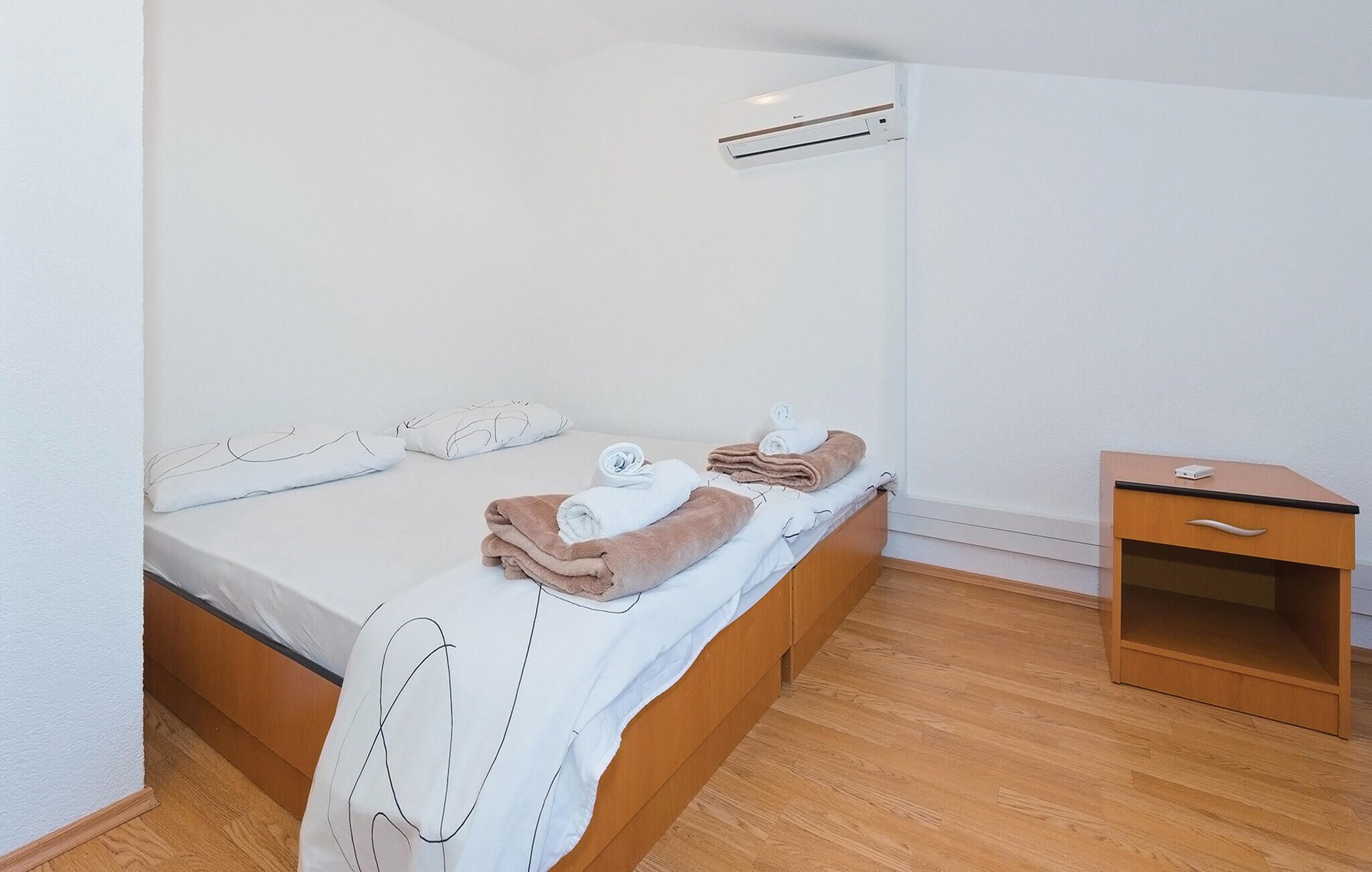 A single bed with a wooden frame, a small bedside table, and an air conditioning unit mounted on the wall.