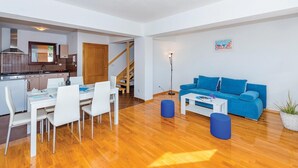 Een moderne woonkamer met een blauwe bank, een witte eettafel en stoelen. Achtergrond toont een keuken met een fornuis en kastjes.
