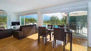 A spacious living area with a sofa, a TV, and a dining table with chairs. There is a large sliding glass door leading to a balcony with a view of the mountains. - Cozy home in Cetina with private swimming pool, can be inside or outside (Cetina)