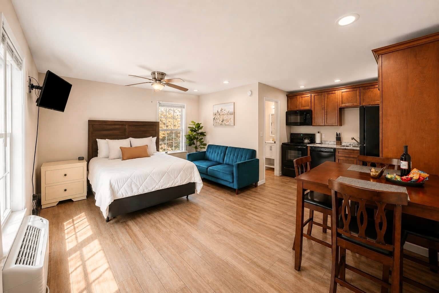 A bedroom with a bed, a blue sofa, a dining table with chairs, a kitchen area with a microwave and stove, and a ceiling fan.