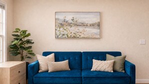 Een blauwe bank met vier kussens, een houten bijzettafeltje en een groot schilderij aan de muur.