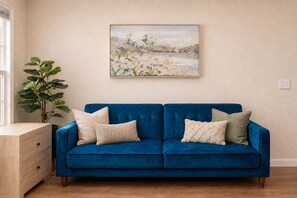 Een blauwe bank met vier kussens, een houten bijzettafeltje en een groot schilderij aan de muur.