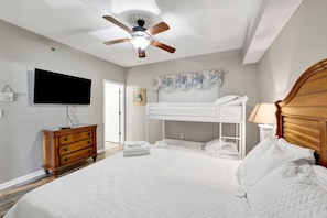 Quarto com cama beliche, ventilador de teto, televisão, cômoda e porta.