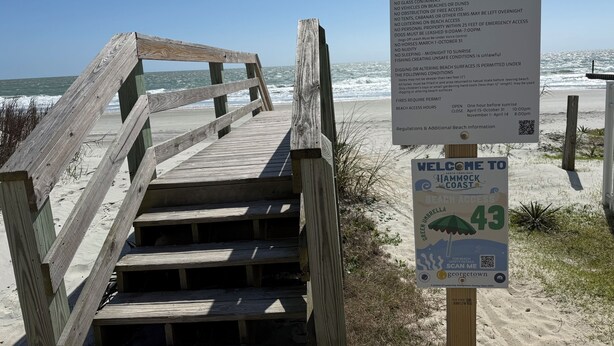 Eine Holztreppe führt zu einem Strand, an dem ein Schild die Strandordnung erklärt und ein Willkommensschild für Hammock Coast zu sehen ist.