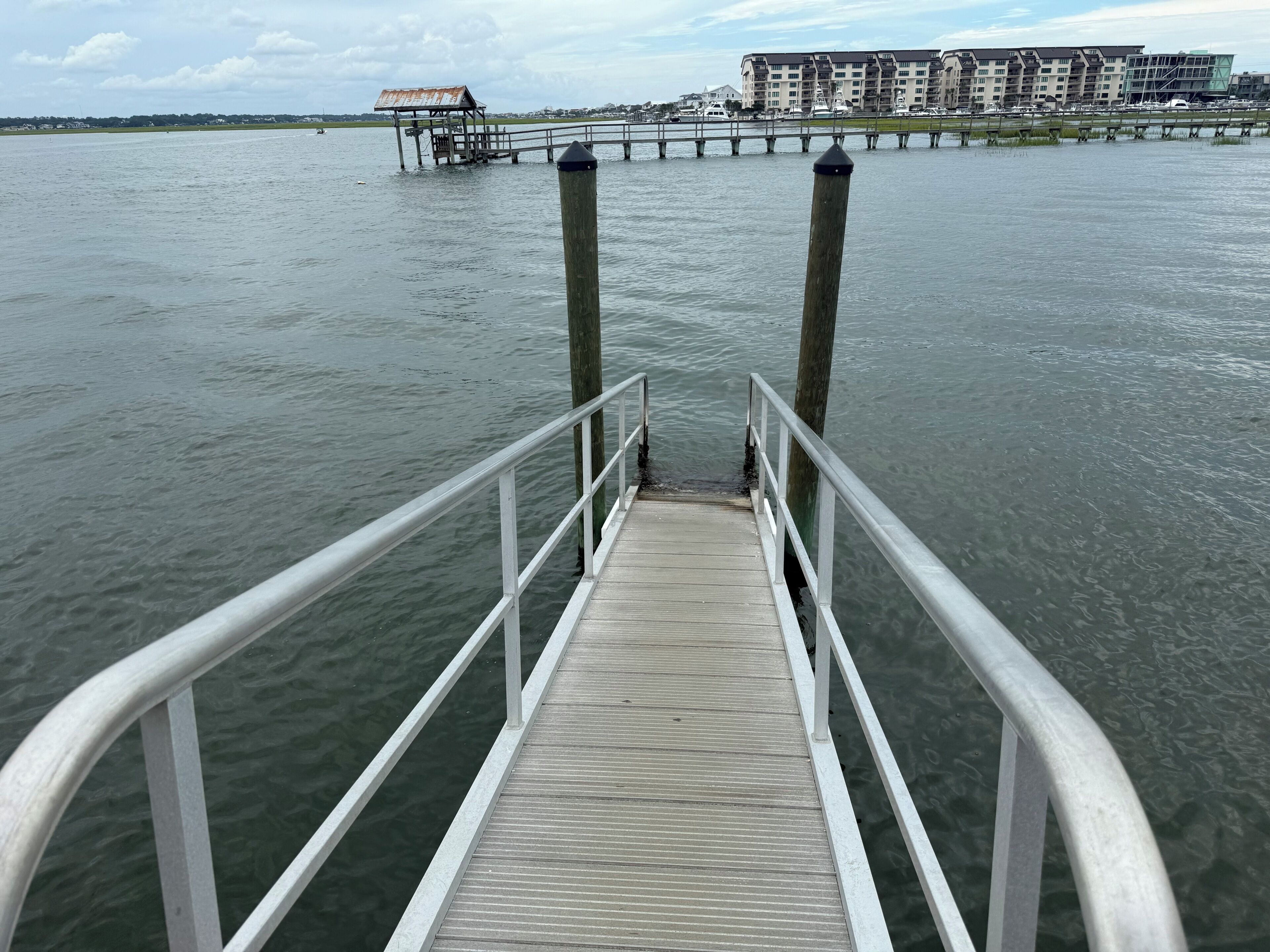 Un muelle de madera que se adentra en aguas tranquilas, con una barandilla de metal, una estructura oxidada a lo lejos y edificios modernos en la orilla opuesta.