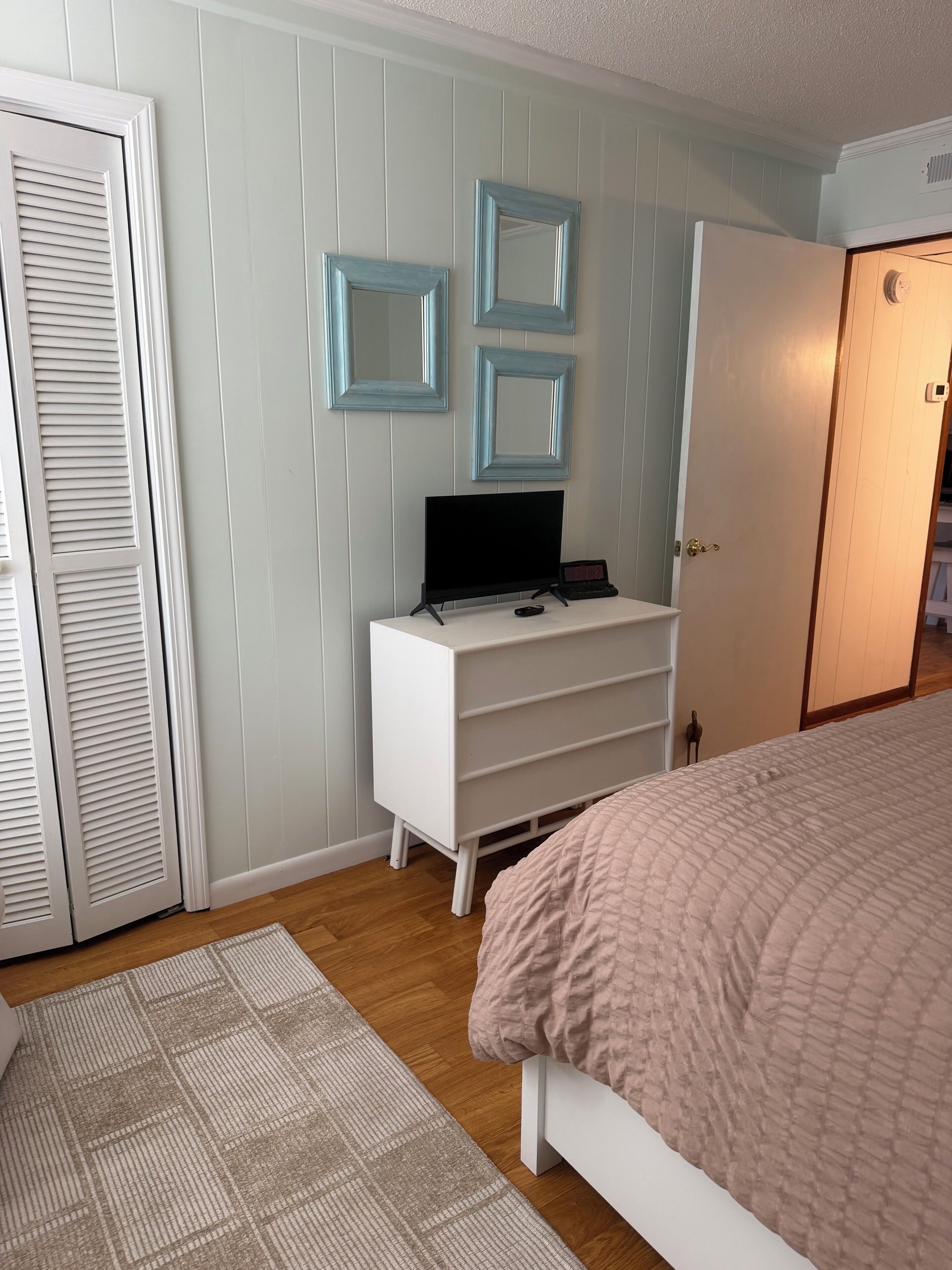 A bedroom with a bed, a TV on a white dresser, a door, and a patterned rug.