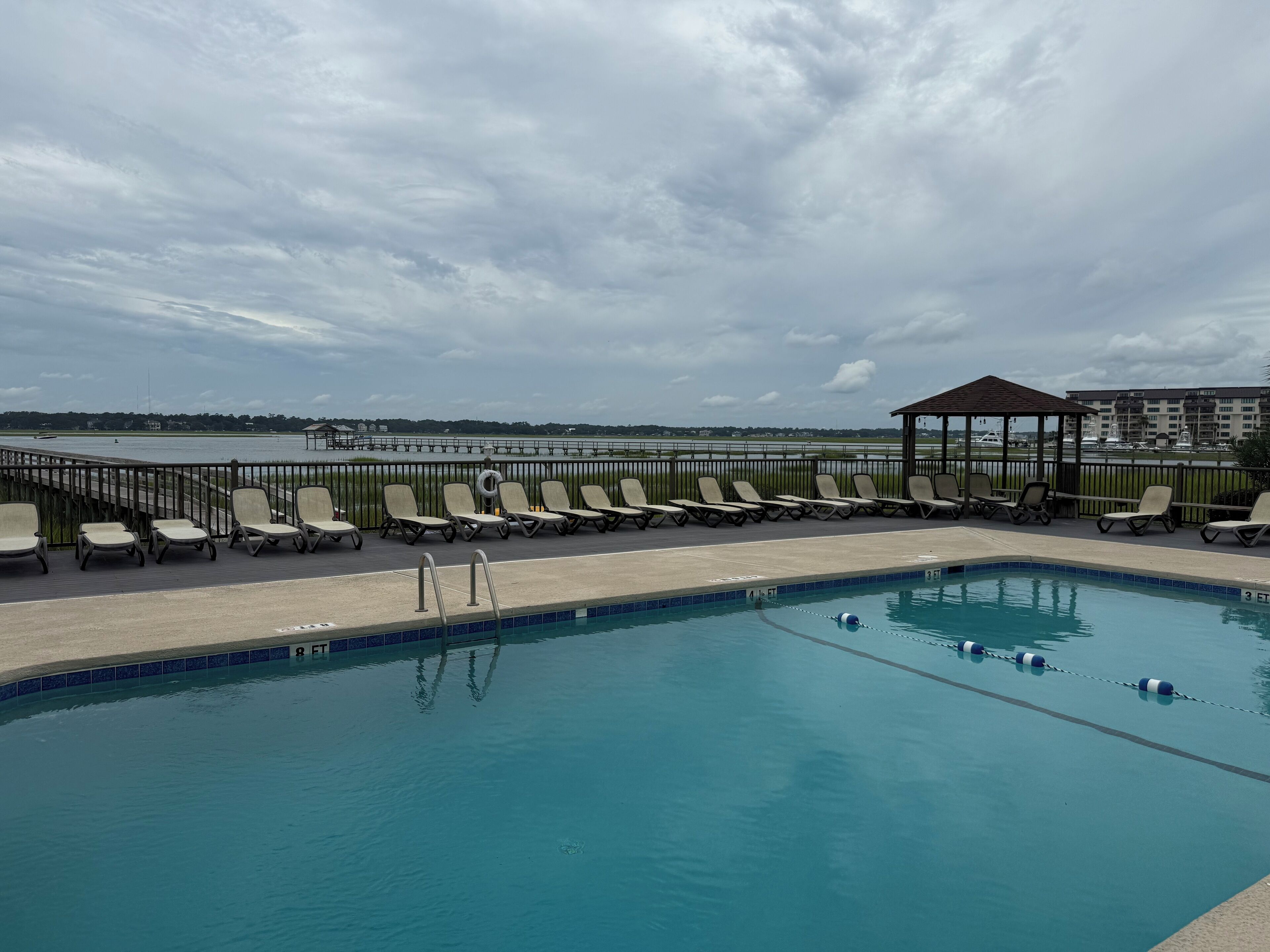 A pool area with lounge chairs and a gazebo, overlooking a body of water and distant buildings.