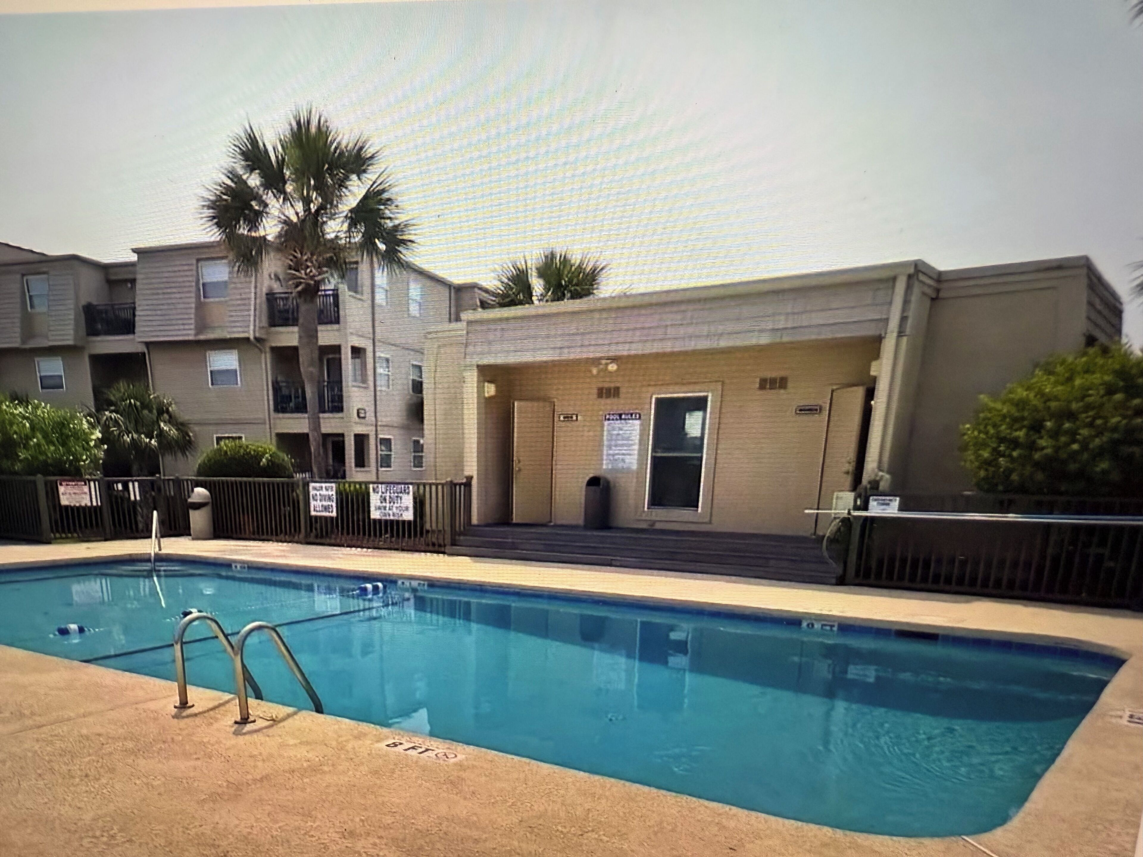 Una piscina con escalera metálica, rodeada por una valla. Se observan palmeras y un edificio con puerta y ventanas al fondo.