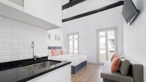 Une cuisine moderne avec un plan de travail noir et des meubles blancs, un lit agrémenté de coussins gris et pêche, un canapé gris avec des coussins pêche, et une grande fenêtre offrant une vue sur les bâtiments.
