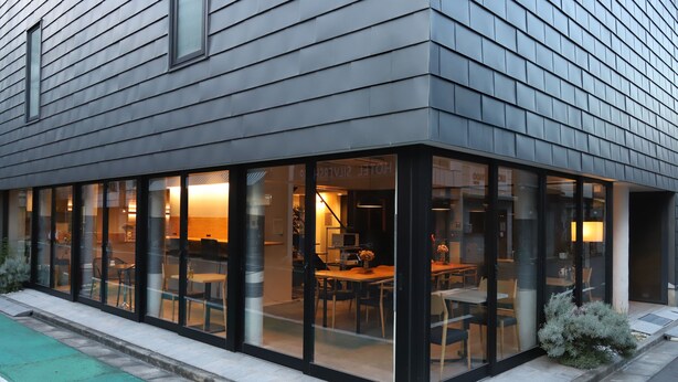 Een modern gebouw met een glazen gevel, een zichtbaar restaurantgedeelte met tafels en stoelen, en een bordje waarop 