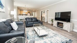 A modern living room with a leather sofa, a glass coffee table, a flat-screen TV, and a kitchen in the background. - Myrtlewood Retreat Near Golf & Ocean Breezes | Magnolia Pointe Luster Leaf 4890-205 (Myrtle Beach)