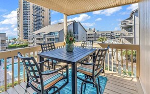 A wooden deck with a table and chairs, overlooking a swimming pool and a view of surrounding buildings. - Ocean Bridge Myrtle Beach Condo (Myrtle Beach)