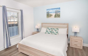 Una camera da letto con un letto, due comodini, una finestra con tende e un quadro a tema spiaggia.