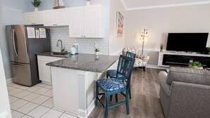 A modern kitchen with white cabinets, a stainless steel refrigerator, and a granite countertop. There is a dining area with a gray sofa and two blue chairs.