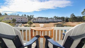 A view from a balcony with two Adirondack chairs and a small wooden table, overlooking a golf course and a row of multi-story buildings with balconies.