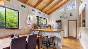 A modern kitchen with wooden beams, a large island with stools, and a view of greenery outside. - Peaceful, creative home in a lush
bush setting, only 300m from the beach! (Skenes Creek)