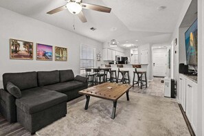 Uma sala de estar moderna com um sofá modular cinza, uma mesa de centro de madeira e um ventilador de teto. Há três quadros nas paredes, uma área de cozinha com armários brancos e uma mesa de jantar com cadeiras.