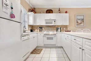 Une cuisine avec des meubles blancs, un four à micro-ondes, une cuisinière et un réfrigérateur. Il y a un tapis sur le sol et un tableau au mur.
