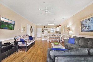 Uma sala de estar espaçosa com um sofá, um sofá cinzento, um espaço de jantar com mesa e cadeiras, um ventilador de teto e uma cozinha ao fundo.