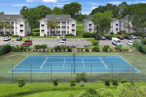 Uma quadra de tênis cercada por um estacionamento com vários carros, e um conjunto de prédios de apartamentos ao fundo.