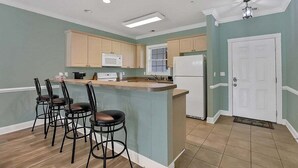 Una cocina con una isla central, taburetes de bar, armarios de madera, un microondas, un refrigerador y una puerta con felpudo.