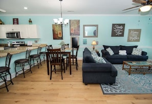Una sala de estar con un sofá azul oscuro, una mesa de centro y un comedor con sillas y una mesa. Hay una zona de cocina con armarios y un microondas.
