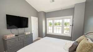 A bedroom with a large bed, a dresser, a flat-screen TV, and a window with a view of greenery.