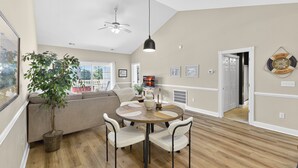 Een moderne woonkamer met een ronde eettafel, comfortabele zitplaatsen, een plafondventilator en een groot raam met uitzicht op het omliggende landschap.