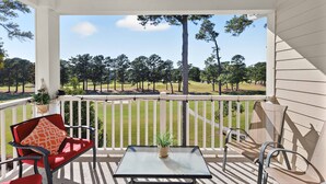 Een overdekt terras met uitzicht op een golfbaan, een tafel met een potplant en twee stoelen met kussens.