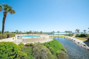 Uma piscina circular com um calçadão ao redor, palmeiras e vista para a praia.