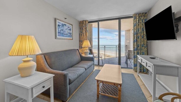 Une chambre d’hôtel moderne avec un canapé gris, une petite table, un téléviseur et un balcon offrant une vue sur la plage.