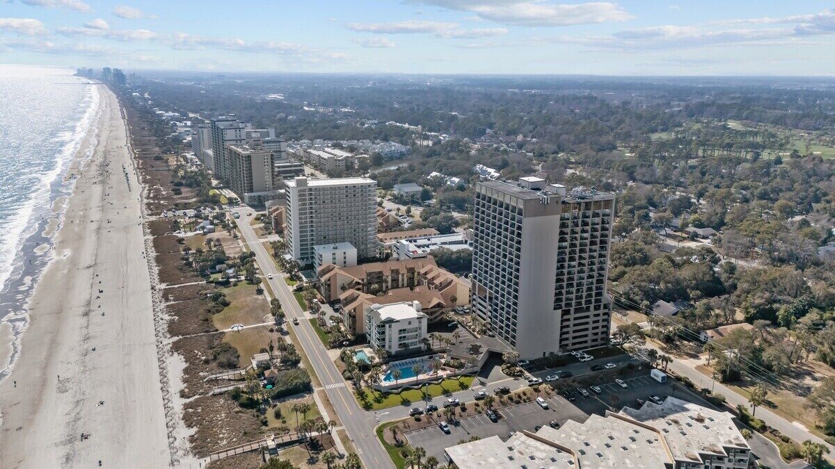 An aerial view of a coastal area with a beach, high-rise buildings, and a golf course.